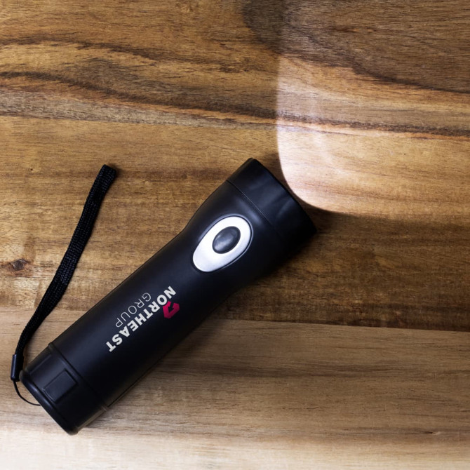 A black rechargeable torch with a silver button, resting on a wooden surface, casting light to one side.