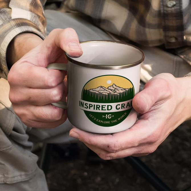 A mug in cream with a gold rim, featuring a nature-themed design and logo on the front.