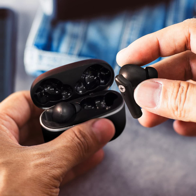 A pair of black earbuds are held above their charging case, with a casual background of denim fabric.