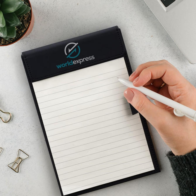 A notepad holder in black with a logo, featuring blank white pages, accompanied by a hand holding a pen, and a plant.