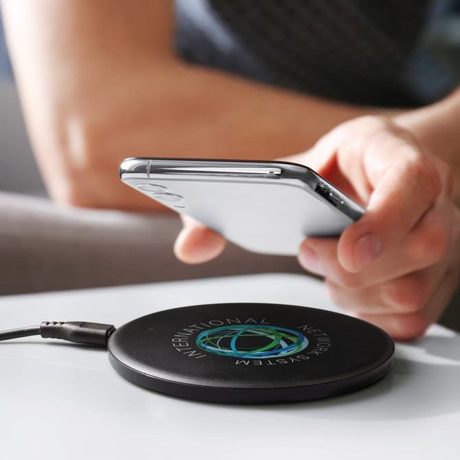 A sleek black wireless charger with a logo, positioned on a table near a smartphone.