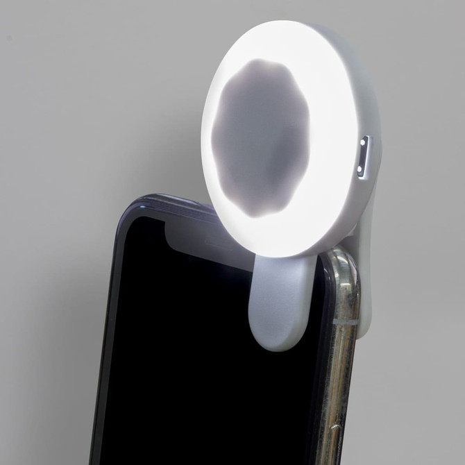 A white halo selfie light attached to a smartphone, illuminating the screen. The product has a circular design.