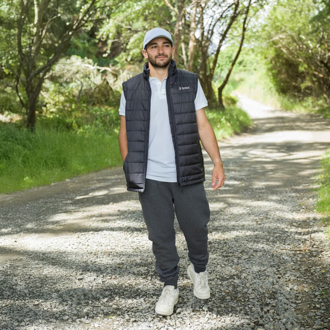 A man wearing a navy puffer vest over a light grey t-shirt and grey joggers, walking along a gravel path in a green setting.