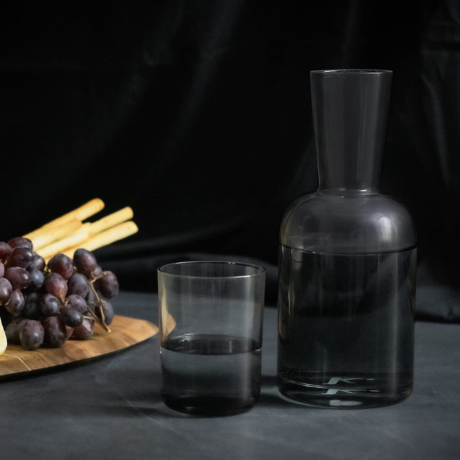 A glass carafe and tumbler set in smoky grey, accompanied by grapes and breadsticks on a wooden platter.