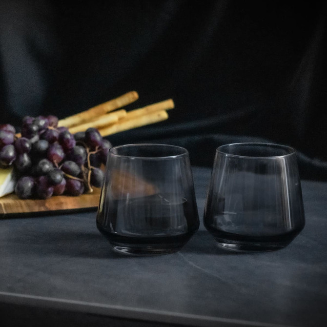 A set of two smoked black whiskey glasses on a grey surface, with grapes and bamboo sticks in the background.