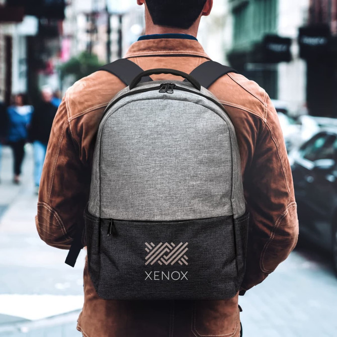 A grey and charcoal backpack with a logo, worn by a person standing on a city street.