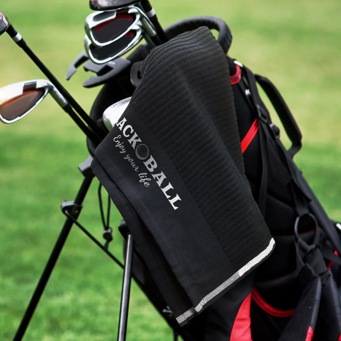 A black golf towel hangs from a golf bag, featuring a textured surface and a logo.