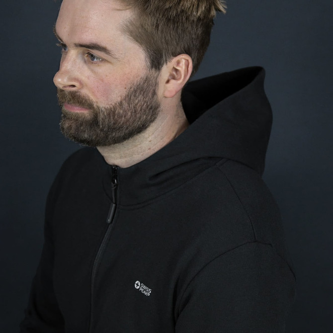 A black hoodie with a zip and a logo, worn by a man with short hair and a beard against a dark background.