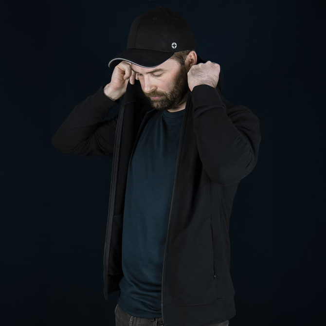 A man in a black Swiss Peak Urban Hoodie adjusts the hood, wearing a black cap. The background is dark.