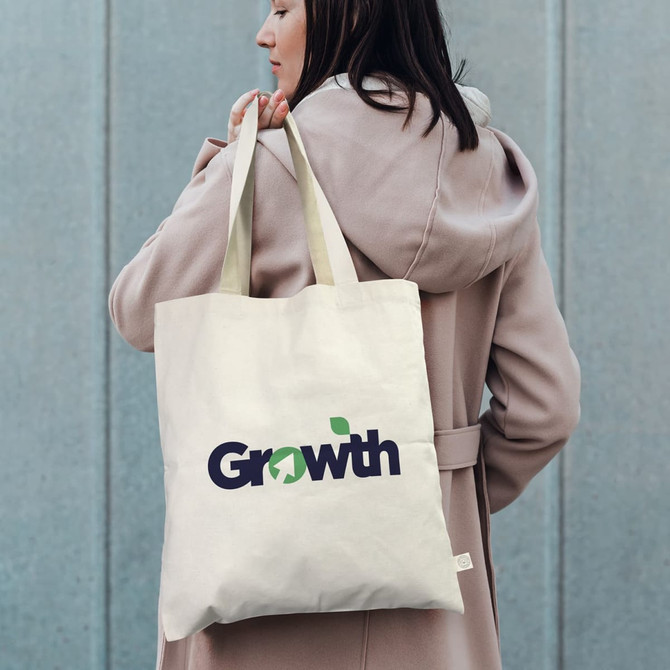 A cream tote bag with sturdy handles, featuring a logo on one side, carried by a person in a light coat.