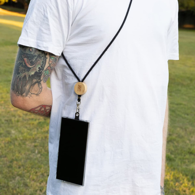 A black phone lanyard hangs from a wooden attachment, worn around the neck of a person in a field.