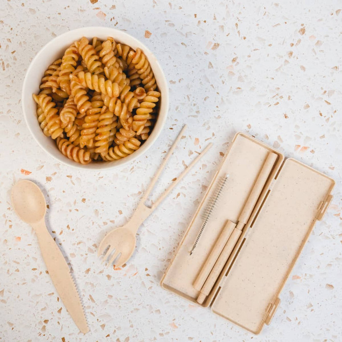 A cutlery set in a beige case, featuring a spoon, fork, and knife, beside a bowl of spiral pasta.