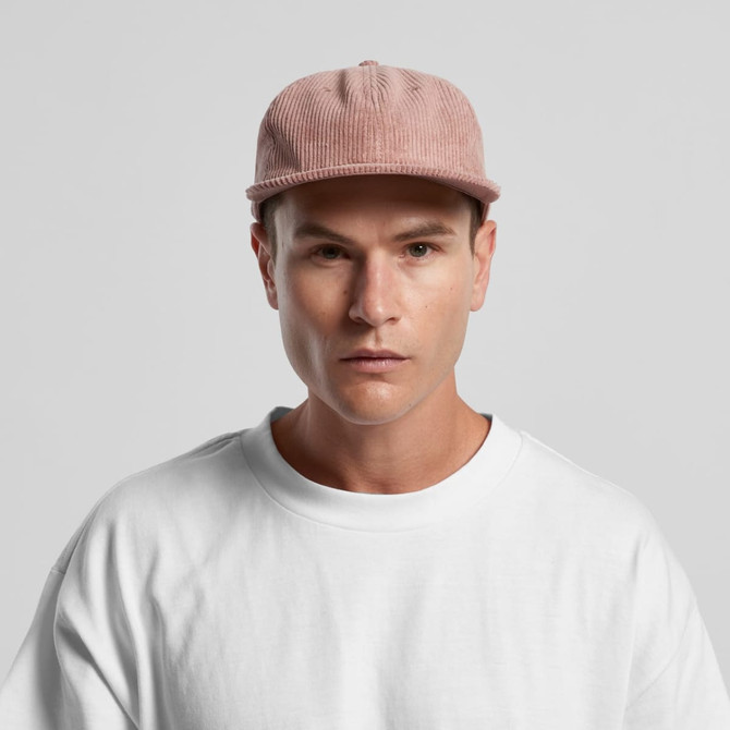 A cord cap in pink is worn by a model in a white t-shirt. The cap features a logo.