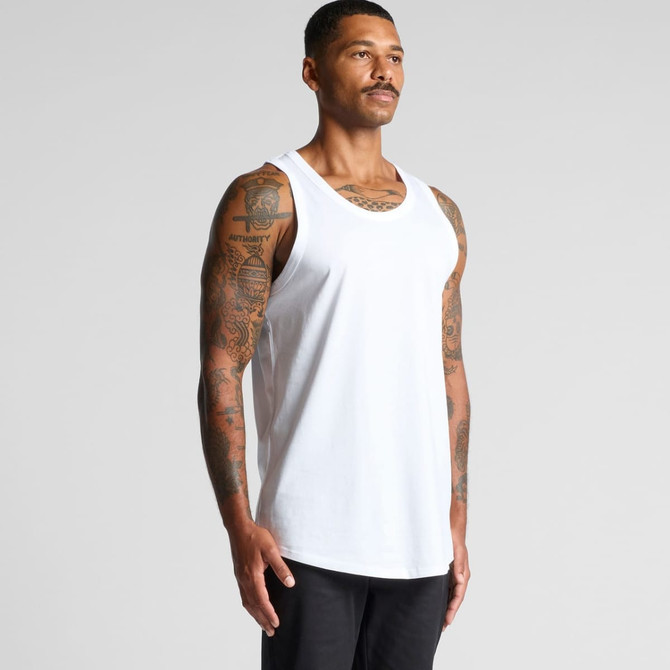 A man stands wearing a plain white singlet, showcasing his tattoos and a branded logo on the garment.