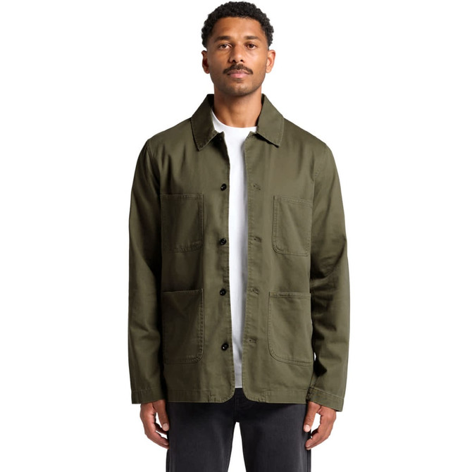A man wearing an olive green Chore Jacket with multiple pockets and a logo. The jacket has a classic collar and button front.