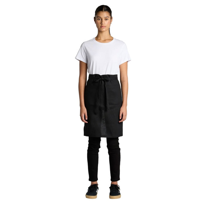 A model wearing a black linen half apron over a white t-shirt and black pants, standing straight with hands at her sides.