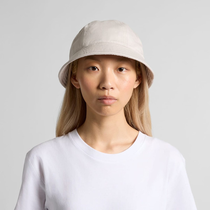 A bucket hat in a light beige colour, featuring a simple design and a logo. The model has long blonde hair.