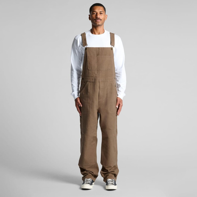Khaki canvas overalls worn by a model, featuring adjustable straps and a relaxed fit. The model pairs them with a white long-sleeve shirt.
