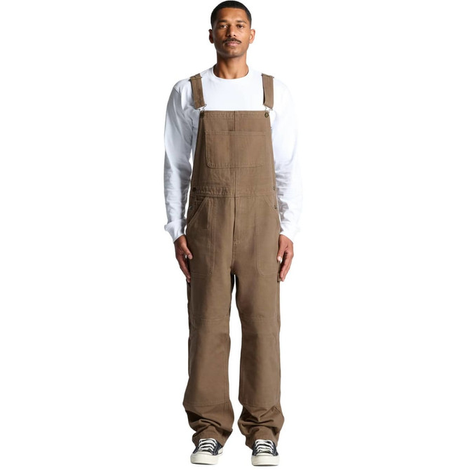 Canvas overalls in brown, featuring adjustable straps and a relaxed fit. Model is wearing them with a white long-sleeve shirt.