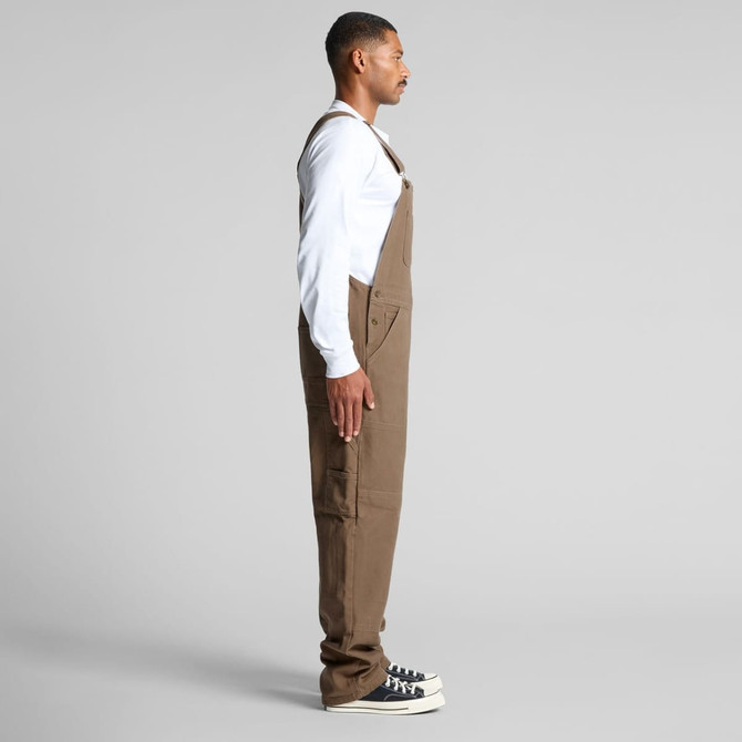 Canvas overalls in brown worn by a model standing sideways, with a white long-sleeve shirt and black shoes.