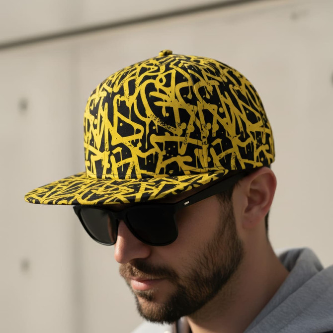A flat peak cap featuring a yellow and black abstract design, worn by a person in sunglasses.