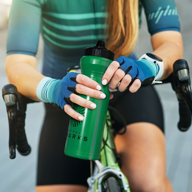 A green drink bottle with a black lid, held by a cyclist wearing blue gloves, sitting on a bicycle.