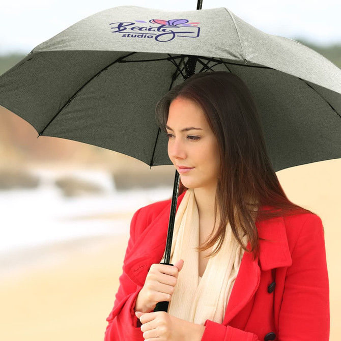 A grey umbrella is held by a woman in a red coat, standing on a beach. The umbrella features a logo.