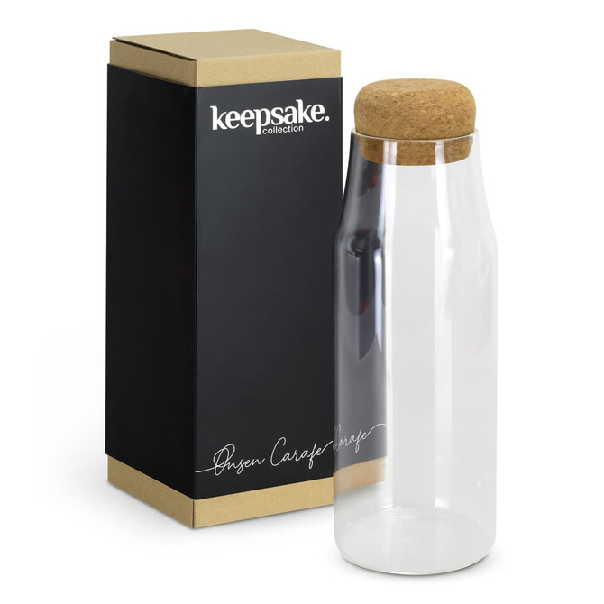 A clear glass carafe with a cork lid, presented beside its black and gold packaging.