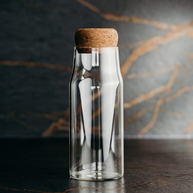 A drink bottle made of clear glass with a cork stopper, set against a textured dark background.