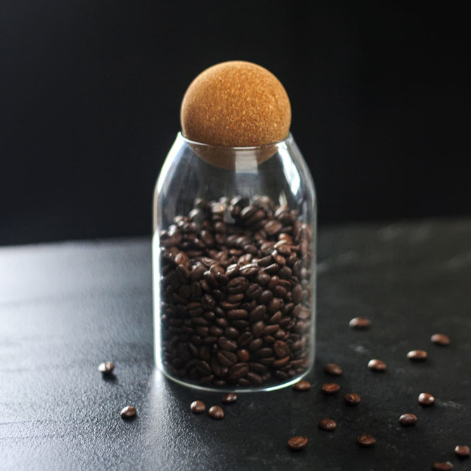 A glass canister filled with coffee beans, topped with a round cork lid, set against a dark background.