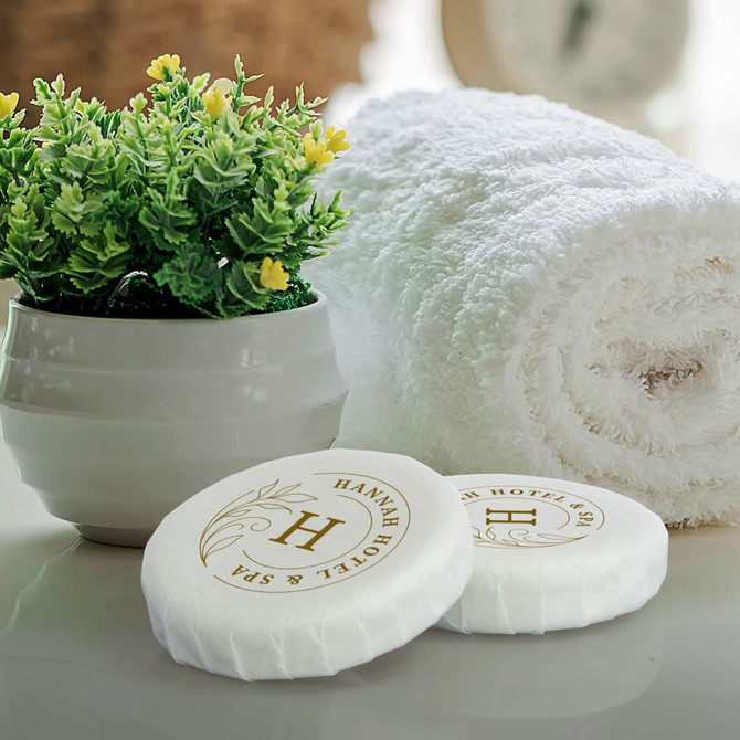 Two white soaps with a logo, placed beside a rolled white towel and a potted plant with yellow flowers.