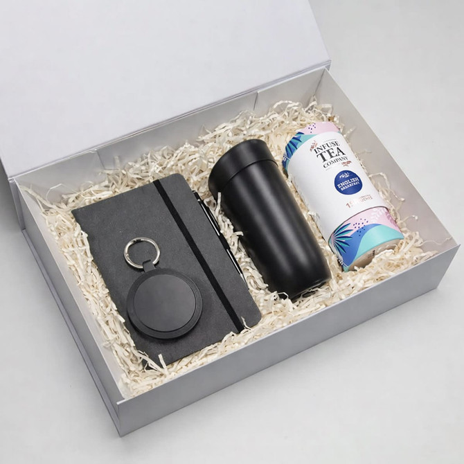 A gift box contains a black drink bottle, a notebook, a keyring, and a canister of tea, all nestled in shredded paper.