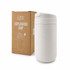 A white coffee cup sits next to its cardboard packaging, featuring a logo and a sleek design.