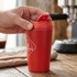 A red coffee cup with a lid, emitting steam, held by a hand on a wooden table. Features a logo.