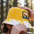 A yellow bucket hat featuring a full-colour patch and various outdoor graphics, held by a person outdoors.