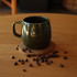 A green ceramic coffee mug sits on a wooden coaster surrounded by coffee beans on a wooden table.