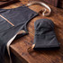 A dark grey canvas apron with light straps and a matching oven mitt, set against a wooden table.