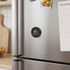 A round black magnetic badge with a logo attached to a stainless steel fridge, alongside a notepad and groceries.