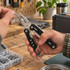 A multi-tool with pliers and a black handle, held in a hand above a wooden workbench with various tools and accessories.