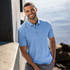 A man stands against a wall, wearing a light blue polo shirt with a collar and buttons, smiling by the water.