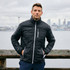 A man stands by a waterfront wearing a black Cutter & Buck Rainier men's jacket with a zippered pocket.