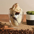 A double wall glass cup filled with a layered dessert topped with whipped cream, beside a small plant and coffee beans.