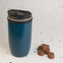 A reusable coffee cup in a teal color with a black lid is positioned beside several chocolate-colored coffee pods.