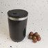 A reusable coffee cup in matte black with a stainless steel band and lid, accompanied by a few chocolate-covered treats.