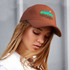 A brown cap with a logo, featuring a colourful mountain design. The cap is worn by a person with long hair.
