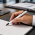A black and white metal pen is held above a notepad, with handwritten notes visible on the page.