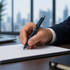 A stylus pen in dark blue, held by a hand, writing on a white sheet of paper with a city skyline in the background.
