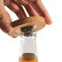 A drink bottle opener made of bamboo is being used to remove a bottle cap, with a clear glass bottle underneath.