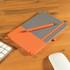 A grey notebook with an orange cover and an orange pen placed on a wooden desk beside a keyboard and mouse.