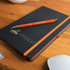 A black notebook with an orange strap and an orange pen resting on top, featuring a logo.